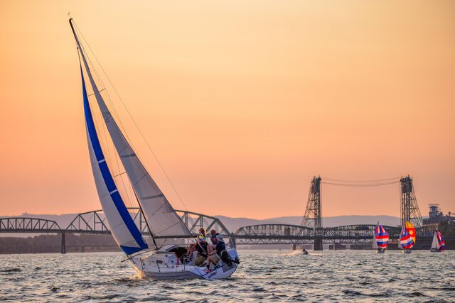 A sloop under blue-and-white sails on a wide river at sunset, with a lift bridge and more sailboats behind it.